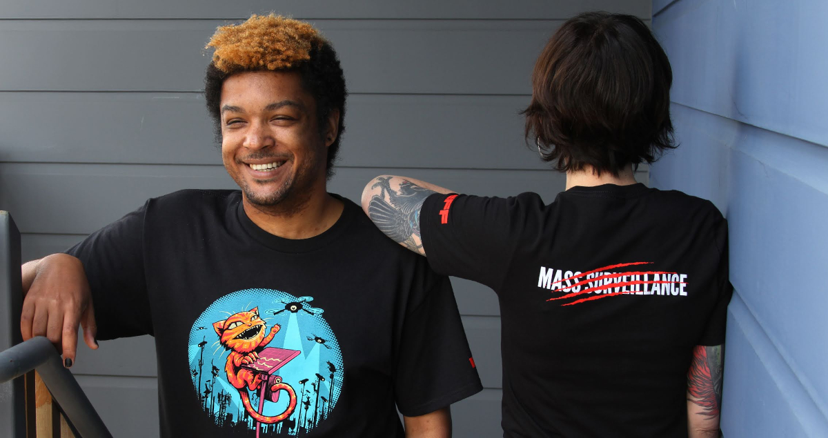Two people wear EFF Claw Back member t-shirts. The front shows a cat swatting at spy cameras and the back says “Mass Surveillance” with red claw marks through it.